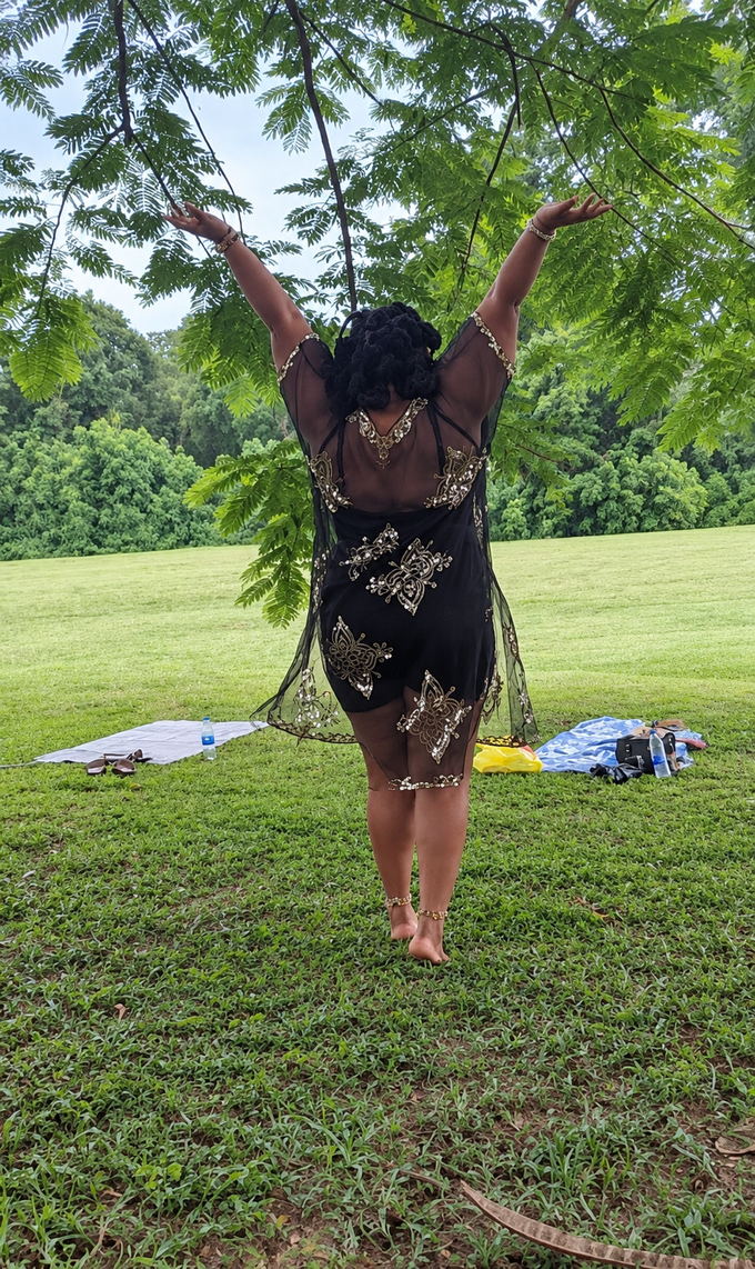 A woman standing on the grass beneath a tree, both arms opened wide to the sky.