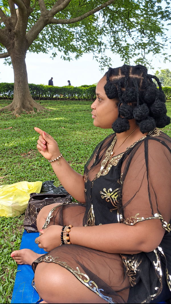 A woman seated cross-legged on a mat on the grass, mid-sentence, gesturing as she speaks.